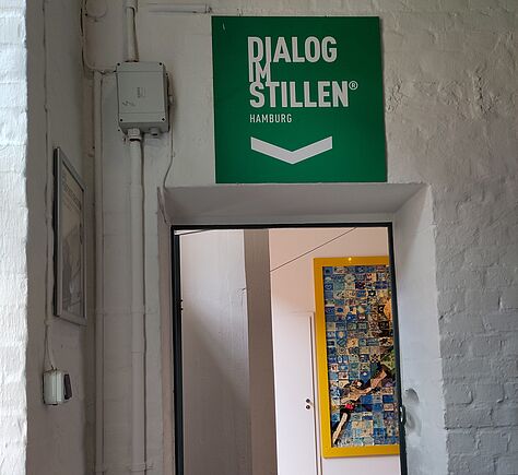 Eine geöffnete Tür führt zum „Dialog im Stillen“, worauf ein grünes Schild über der Tür hinweist. In dem Raum sind Leitstreifen auf den Holzboden. Im Hintergrund ist ein großes gelbgerahmtes Bild.