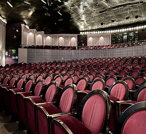 Eine Gesamtansicht von dem Theatersaal. Die Stühle haben eine ovale Rückenlehne sind aus Holz und rot bezogen. An der Saaldecke sind viele Lampen und Scheinwerfer zu sehen. Im Hintergrund ist auch der erste Rang zu erkennen.