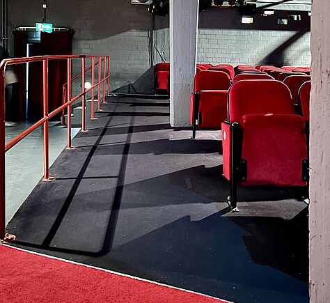 Teilansicht des Kinosaals mit mehreren Reihen roter Sitze. Auf der linken Seite ein Gang mit rotem Teppich. Ein rotes Geländer grenzt den schräg aufsteigenden Gang vom Raum ab. Die Wände sind grau, es gibt sichtbare Lüftungsrohre.