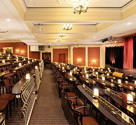 Ein festlich beleuchteter Theatersaal mit klassischer Deckengestaltung und warmen Wandfarben. Reihenweise stehen schmale Tische mit gepolsterten Stühlen, auf jedem Tisch leuchtet eine kleine Lampe mit Schirm. In der Mitte verläuft ein mit gemustertem Teppich ausgelegter Gang, links ist ein Geländer aus hellen, gebogenen Metallstreben. Die Tischreihen sind links aufsteigend und rechts zur Bühne hin abfallend, von der ein Vorhang hängt. Die Atmosphäre wirkt elegant und einladend.
