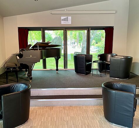 Bühne mit schwarzem Flügel und Sitzgruppe. In einem taghellen Raum steht auf einer kleinen erhöhten Bühne links ein schwarzer Flügel der Marke "Steinway & Sons". Rechts auf der Bühne sind drei schwarze, lederne Sessel kreisförmig um einen kleinen runden Tisch angeordnet. Vor der Bühne stehen auf dem Parkettboden zwei weitere schwarze Ledersessel. Im Hintergrund sieht man durch große Fenster Grünflächen und Bäume.