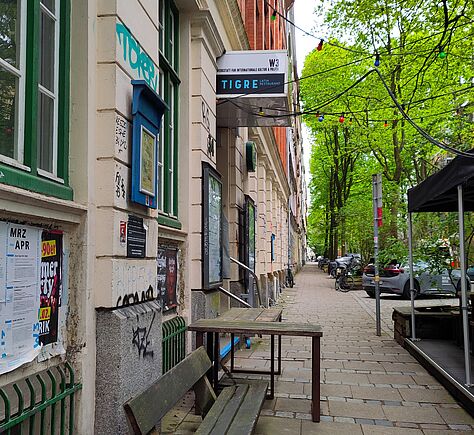 Blick auf eine gepflasterte Straße mit einer grünen Holzbank und einem Schild mit der Aufschrift 'W3´und `TIGRE' an einem Backsteingebäude, umgeben von Bäumen und geparkten Autos.