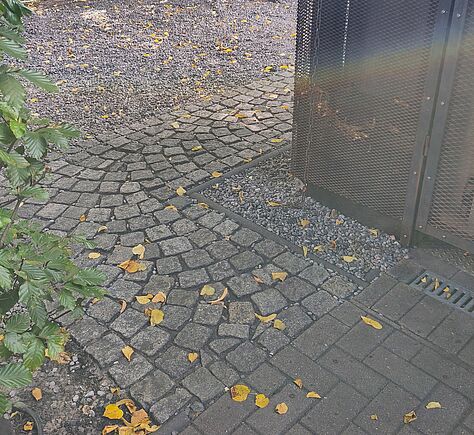 Ein schmaler gepflasterter Weg führt nach rechts um die Ecke eines Gebäudes, das mit einem Metallgitter verkleidet ist. Rechts und links von dem Weg liegt grauer Kies. Auf dem Boden liegen verstreut gelbe Blätter. Links ragt ein belaubter Strauch ins Bild. Hinter einem Zaun befindet sich eine Straße und Häuser.
