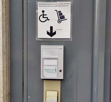 Eine graue Wand mit einer Gegensprechanlage und einem großen Klingelknopf darunter. Darüber hängt ein Schild mit Symbolen für Rollstuhl und Transport sowie einem Pfeil nach unten. Die Oberfläche wirkt glatt und funktional.