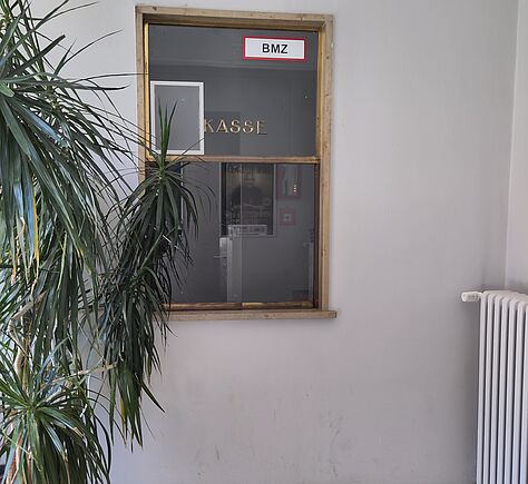 Kleiner Kassenbereich mit hellem Fliesenboden und weißer Wand. Das Kassenfenster hat einen Holzrahmen und dunkles Glas, links steht eine große Pflanze, rechts ein weißer Heizkörper.