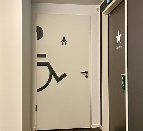 Blick auf die weiße WC-Tür. Darauf im Anschnitt ein großes Rollstuhl-Symbol und mittig ein Icon für Wickeltisch. An der rechten Wand eine zweite WC-Tür mit der Aufschrift: Künstler