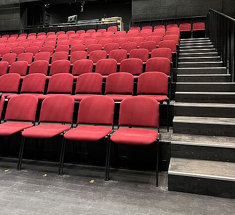Blick in den leeren Theatersaal mit vielen roten Sitzen in aufsteigenden Reihen. Auf der rechten Seite ein Gang mit schwarzen Stufen. Im Hintergrund ein Regiepult und Scheinwerfer.