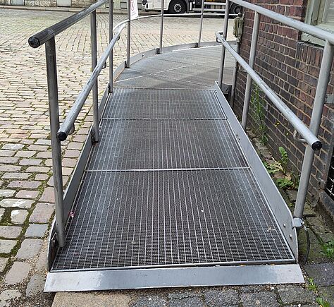 Auf dem Gehweg mit Kopfsteinpflaster befindet sich eine breite Metallrampe um die Ecke an der Hauswand. Rechts und links der Rampe sind zwei Handläufe, ein niedriger und ein höherer. 