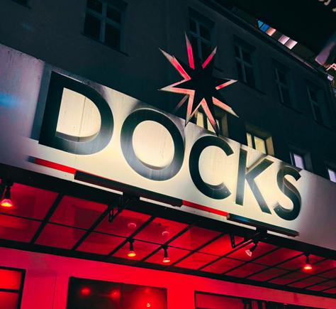 Großes, beleuchtetes Schild mit dem Schriftzug 'DOCKS' in serifenlosen Großbuchstaben. Über dem Schriftzug befindet sich ein stilisierter Stern mit acht Zacken. Das Schild ist an der Fassade des Gebäudes angebracht. Der Bereich unter dem Schild ist in rotes Licht getaucht.