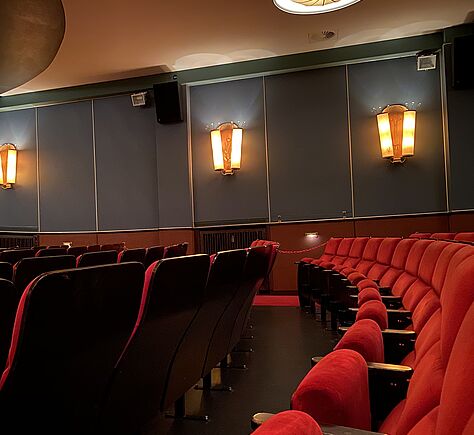 Es sind zwei Sitzreihen zu sehen, rote Kinosessel, viel Bewegungsfreiheit zwischen den Reihen. Im Hintergrund Wandlampen.