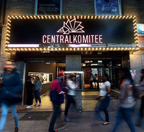 Leuchtreklame mit der Aufschrift „CENTRALKOMITEE“ an einer Gebäudefassade am Abend. Über der Schrift ist ein stilisiertes, fächerförmiges Logo. Rund um das schwarze Schild verläuft ein Rahmen aus vielen kleinen gelblichen Glühbirnen, die leuchten. Darunter der Eingangsbereich. Vor dem Eingang stehen und gehen mehrere Personen; einige sind scharf, andere durch Bewegung verwischt. Davor der Gehweg. 