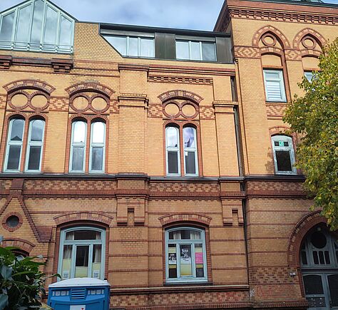 Fassadenansicht eines historischen Gebäudes aus gelbem und rotem Backstein. Die Fassade zeigt viele dekorative Rundbögen und Klinkerverzierungen über Fenstern. Mehrere hohe Fenster mit weißen Rahmen, in einige spiegelt sich der Himmel. Im unteren Bereich sind zwei große Fenster sichtbar, davor steht unten links eine blaue mobile Rolli-Toilette. Am rechten Bildrand ragt ein Baum mit grün-gelbem Laub hervor. Links am Rand wachsen einige Sträucher. Die Tür ganz rechts (der Haupteingang) hat ein Bogenfenster darüber und trägt die Hausnummer 79. Der Himmel ist blau mit einigen Wolken.