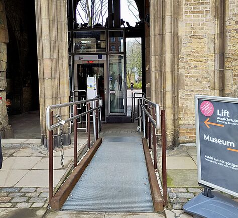 Eine Metallrampe mit Geländern führt zu einem gläsernen Aufzug in einer Stein- und Backsteinumgebung, rechts steht ein graues Schild mit orangefarbenem Pfeil zu „Lift“ und „Museum“.