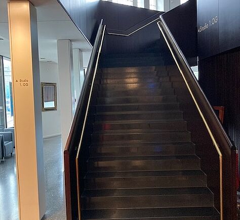 Eine breite Treppe mit kontratsreich abgesetztem Handlauf links und rechts. Erste und letzte Stufe sind nicht markiert. Ganz oben macht die Treppe eine Rechtskurve.