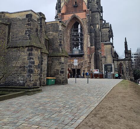 Ein historisches Gebäude aus hellem und dunklem Stein mit einem hohen Turm und großen Bögen. Vor dem Eingang liegt ein gepflasterter Weg, der leicht ansteigt. Am Eingang sind mehrere Personen zu sehen.