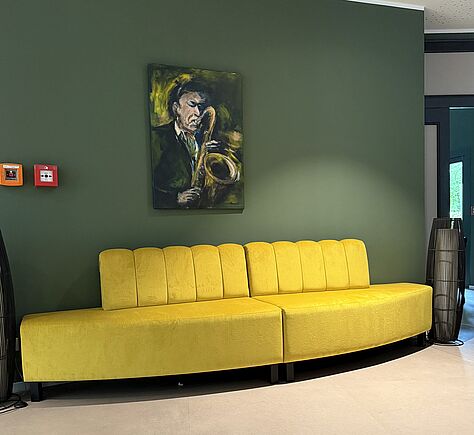 Vor einer nach außen gewölbten grünen Wand steht ein gelbes 2-teiliges Sofa. Darüber hängt ein Gemälde: Portrait eines Saxophonspielers mit seinem Instrument vor grün gehaltenem Hintergrund 