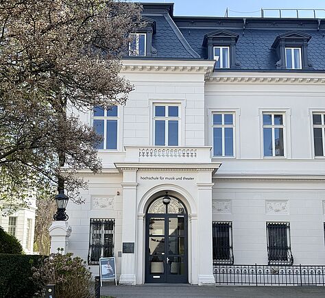 Die Fassade eines historischen, weißen Gebäudes mit einem dunklen Dach und mehreren Fenstern. Über dem Eingang die Aufschrift: Hochschule für Musik und Theater. Die Eingangstür ist aus Glas, groß und mit einem Bogen darüber. Links daneben steht eine Laterne auf einer Säule. Der Himmel ist blau.