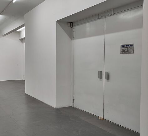 Weiße Doppeltür in einem weißen Flur mit grauem Boden, an der Tür ein kleines Schild mit schwarzer Schrift