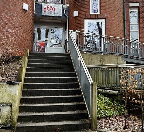 15 dunkle Stufen führen zu einer Glastür; in den Scheiben spiegeln sich kahle Baumäste. Über dem Eingang hängt ein Schild mit dem Wort „ella“ in roter Schreibschrift. Links neben der Tür steht ein Werbeaufsteller mit einem Foto. Rechts verläuft ist der Handlauf der Rampe erkennbar.