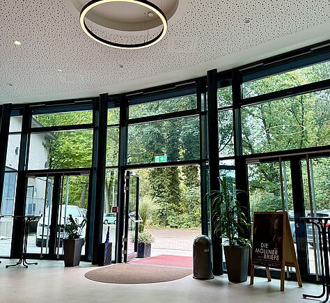 eine große bodentiefe Fensterfront mit schwarzen Rahmen, in der Mitte ist die Eingangstür göffnet. Im Eingang liegen sowohl draußen wie drinnen jeweils eine Schmutzmatte in Rottönen. An der hellen Decke hängt eine runde leuchtstoffröhre.