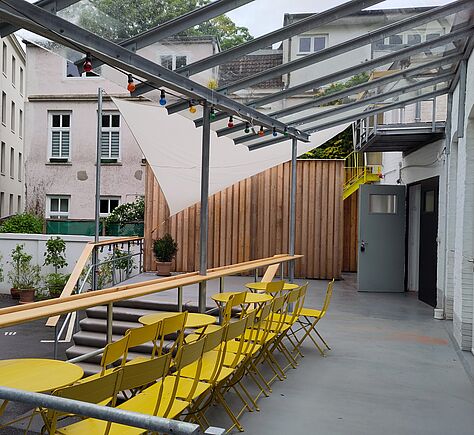 In einem Innenhof zwischen Wohnhäusern ist ein Podest zu sehen, zu dem eine Treppe führt. Im hinteren Teil durch ein Segel, im vorderen durch eine transparente Überdachung geschützt. Die aus Rohren gebauten Geländer sind oben auf mit breiten hellen Holzleisten versehen.