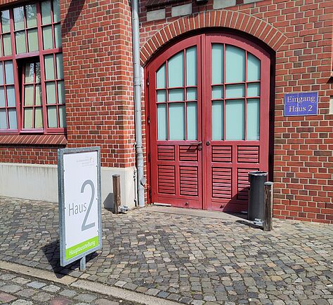 Eine rote Doppeltür aus Holz mit Glaselementen prägt den Eingang eines Backsteingebäudes. Rechts neben der Tür befindet sich ein blaues Schild mit weißer Schrift „Eingang Haus 2“. Im Vordergrund steht ein großes Schild mit der Aufschrift 'Haus 2' auf einem gepflasterten Weg.
