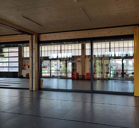 Der Holzbau des Saales mit der breiten Schiebetür und gelben Vorhängen. An der Außenwand zum Hof hin stehen mittig 2 rote Sessel mit einem Couchtisch dazwischen. Links im Bild ist der Teil eines weißen Tresens zu sehen.