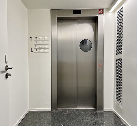 Frontaler Blick auf den aluminiumfarbenen, den Fahrstuhl. Links hat er ein Fenster in Form eines Bullauges
