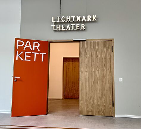 Der linke Türflügel zum Theatersaal steht offen. Darauf in großen weißen Buchstaben das Wort Parkett. Über der Tür in weißen Buchstaben. Unten im Bild ist ein Teil des Boden Leitsystems zu sehen.