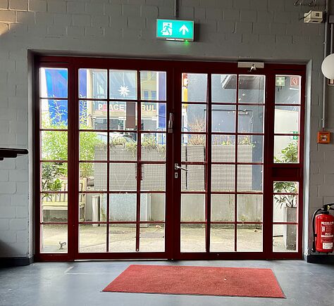 Vom Foyer aus gesehen, eine große doppelflügelige Tür mit rotem Rahmen und vielen kleinen Fensterscheiben. Über der Tür ein grünes Notausgang Schild. An der Wand rechts hängt ein Feuerlöscher. Auf dem Boden vor der Tür liegt eine rote Fußmatte.
