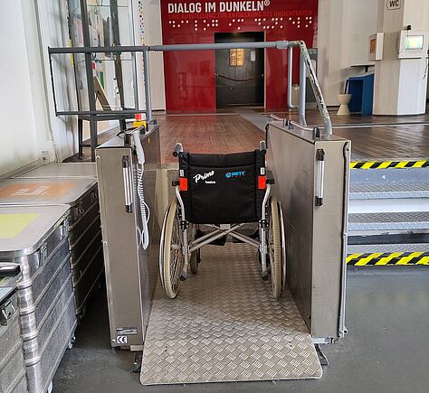 Der Hebelifter links neben einer Treppe überwindet vier Stufen. Die untere und obere Stufe ist gelbschwarz gestreift markiert. Auf dem Lifter steht ein einfacher Rollstuhl. Eine rot gerahmte Tür befindet sich im Hintergrund, da steht: DIALO IM DUNKELN.