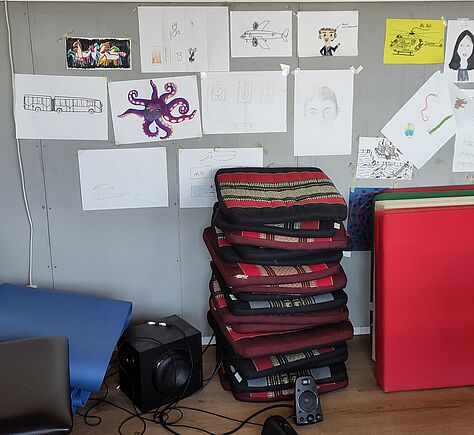 An der Leichtbauwand liegen und stehen (v.l.n.r.) eine blaue Gymnastikmatte, ein Subwoofer mit kleinen Lautsprechern, ein Stapel Sitzpolster, feste bunte Turnmatten. An der Wand hängen selbstgemalte Bilder auf Papier