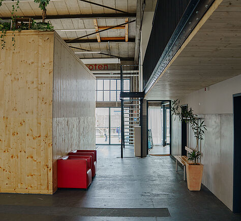 Der Gang liegt zwischen der hölzernen Außenwand des Tanzsaales und der Wand, in der die Türen zu den WC's abgehen. Links steht ein flaches rotes Sofa. Im Hintergrund große Glasfenster mit Blick auf das Außengelände