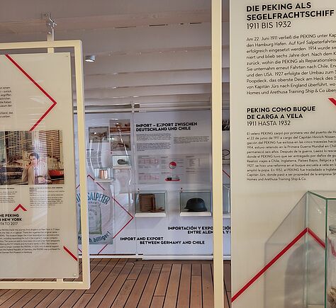 Blick in eine Ausstellungsfläche mit hellem Holzboden und weißen Wänden. Zwei große Informationstafeln hängen frei im Raum. Die Texte sind auf deutsch, spanisch und englisch. Es geht um die Peking als Segelfrachtschiff und Filmkulisse. Im Hintergrund sind Vitrinen mit Ausstellungsstücken und weitere Schautafeln zu sehen. Die Gestaltung wirkt modern und übersichtlich.
