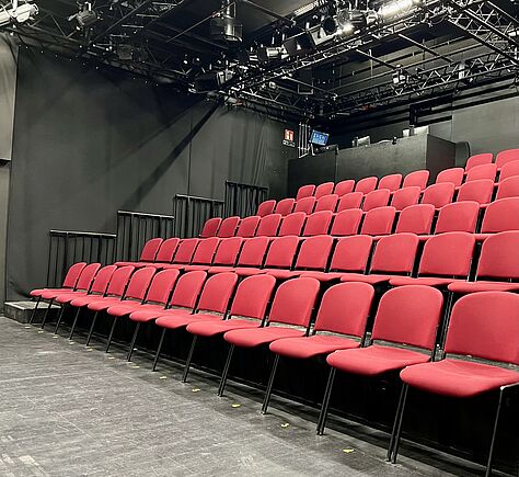 Blick in den leeren Theatersaal mit vielen roten Sitzen in aufsteigenden Reihen. Im Hintergrund Scheinwerfer.