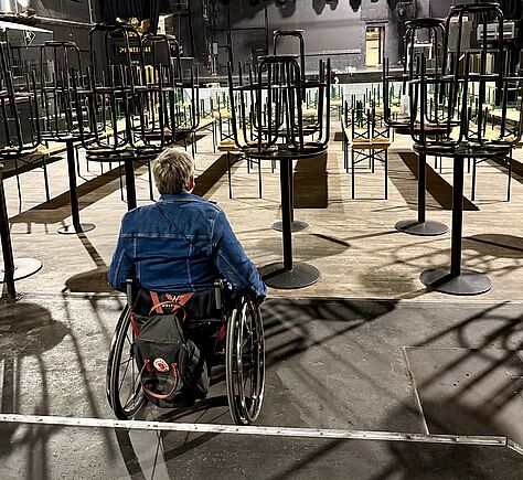 Eine Person im Rollstuhl sitzt mit dem Rücken zur Kamera auf einem erhöhten Podest im Docks. Im Saal stehen zahlreiche umgedrehte Stühle auf Tischen. Der Saal ist leer, die Beleuchtung ist gedämpft. Im Hintergrund sind technische Geräte und Bühnenausstattung zu erkennen.