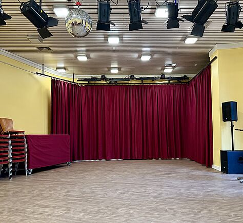 Der leere Saal an der Rückwand ein dunkelroter Vorhang. Der Boden ist mit hellem Laminat belegt. An der Decke hängen mehrere schwarze Scheinwerfer im Vordergrund und eine silberne Discokugel. Weiter hinten mehrere rechteckige eingeschaltete Deckenleuchten, Links stehen gestapelte Stühle auf einem Rollwagen und ein länglicher Tisch auf Rollen, beides mit weinroten Tüchern abgedeckt. Rechts steht eine schwarze Lautsprecherbox auf einem Stativ und eine andere am Boden. Die Seitenwände sind hellgelb gestrichen.