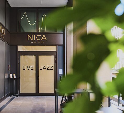 Im Vordergrund verdecken unscharfe grüne Blätter einen Teil des Bildes. Dahinter befindet sich der Eingang. Über der Tür mit dunklem Rahmen und hellen Glasflächen steht in goldenen Buchstaben der Name »NICA JAZZ CLUB«. Auf den beiden Türflügeln ist jeweils das Wort »LIVE« und »JAZZ« in dunklen Großbuchstaben. Links neben der Tür steht ein kleiner Tisch mit Flyern im Ständer.