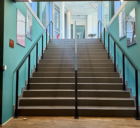 Eine breite Treppe mit Handläufen links und rechts, die in der Mitte auch mit einem Handlauf voneinander getrennt sind. Die Stufen sind dunkel und aus Holz. Die Wände sind Türkisblau. Oben auf der Treppe sind Fenster. 