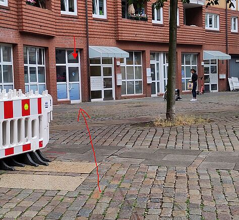 Blick auf einen Platz mit altem Kopfsteinpflaster. Im Hintergrund eine moderne, niedrige Häserfassade mit Sprossenfenstern.
