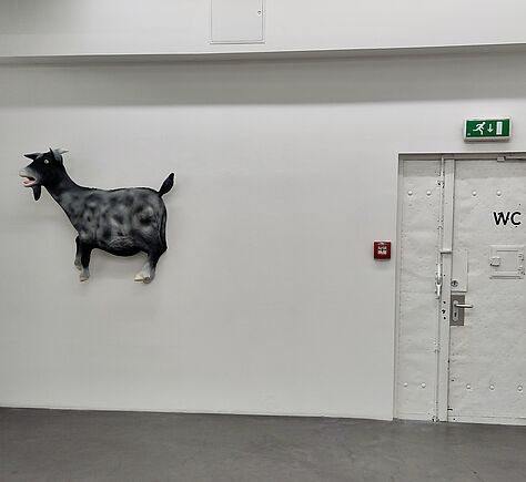 Rechts im Bild eine weiß gestrichene Brandschutztür mit schwarzer Beschriftung "WC". Darüber ein grünes Leuchtzeichen mit dem Piktogramm für Fluchtweg. Links neben der Tür ein rotes Kästchen mit Fenster für den Feueralarm. An der weißen Wand links neben der Tür hängt ein schwarzes Relief einer Ziege in Lebensgröße. Sie hat das Maul halb geöffnet. Im Vordergrund grauer Bodenbelag.