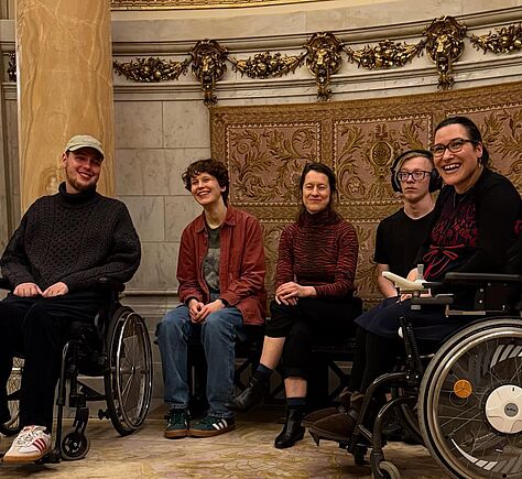 Fünf Personen posieren gemeinsam in einem prunkvollen Raum mit marmorner Säule und aufwendig verziertem Wandteppich und Gemälde im Hintergrund. Zwei der Personen sitzen in Rollstühlen, die anderen drei auf Bänken dazwischen. Alle sind entspannt und lächeln. Die Atmosphäre wirkt freundlich und harmonisch. Das Dekor ist sehr kunstvoll mit goldenen Verzierungen und klassischen Motiven.