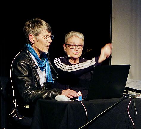 Karen Müller und Kerstin Hagemann sitzen vor einem Laptop bei einer Präsentation. Die Umgebung ist dunkel, beide tragen ein Mikrophon. 