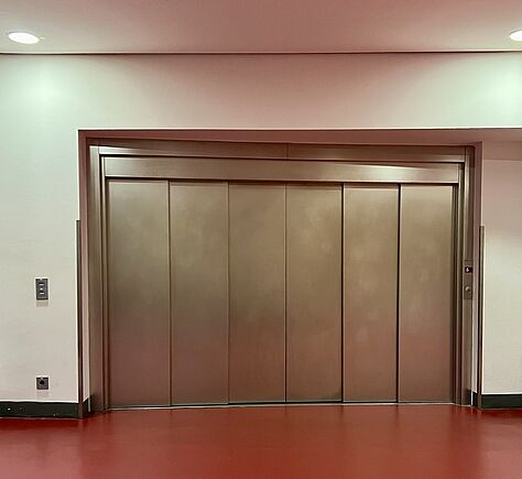 Der große Fahrstuhl mit geschlossenen Türen. Der Fahrstuhl ist aluminiumfarben, links neben den Türen ist ein Bedienfeld mit Knöpfen. Der Boden ist rot und die Wände sind weiß.