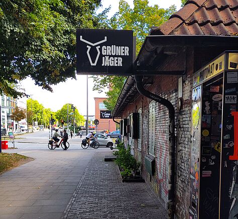 Ein breiter Gehweg, teils Kopfsteinplaster, Backsteingebäude rechts, an dem ein schwarzes Schild mit weißem Logo und Schriftzug »GRÜNER JÄGER« hängt, links ragt ein großer Baum ins Bild, urbane Straßenszene im Hintergrund