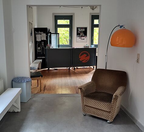 Heller Raum mit unterschiedlichen Sitzmöglichkeiten: Im Vordergrund rechts steht ein gemusterter, brauner Sessel mit Rollen nahe einer Wand,  eine orange Steh-Bogenlampe daneben. Auf der linken Seite eine schlichte weiße Sitzbank vor einem Heizkörper, davor ein kleiner Hocker mit gemustertem Kissen. Im hinteren Raum steht eine Theke mit dem orangenen monsun.theater-Logo. Dahinter ein schwarzer Kühlschrank mit mehreren Aushängen und links an der Wand davor eine weitere weiße Bank. Zwei große Fenster lassen Tageslicht herein, Zwischen denen ein Poster hängt.
