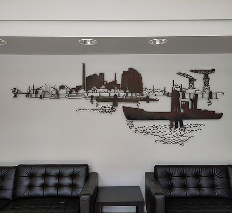 Im Foyer stehen schwarze Ledersofas und ein kleiner Tisch vor einer weißen Wand. Darüber hängt ein großes Metallbild mit Hafenmotiven, Schiffen und Kränen in dunklen Rostfarben.