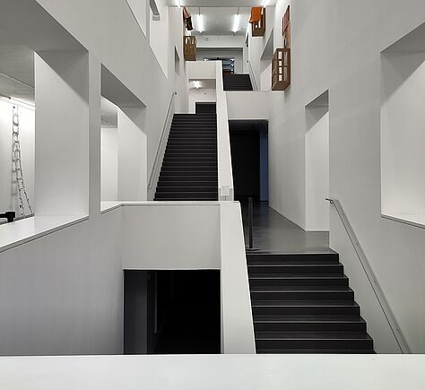 In einem lichten Schacht sind die Treppen mit dunklen Stufen jeweils abwechselnd pro Stockwerk rechts und links an die Wand gebaut. Zur Mitte hin sind die Treppen mit einer durchgängigen Mauer gesichert. Die Wände sind weiß, minimalistische Architektur.