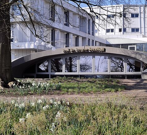 Das Bild zeigt die Fassade der JazzHall. Der Name steht in goldfarbenen Buchstaben über den Fenstern: Prof. Dr. Langner Jazz Hall. Das Gebäude ist niedrig und hat eine große, leicht bogenförmige Glasfront. Vor den Fenstern ist eine Wiese mit vielen weißen Blüten. Rechts und links Bäume ohne Blätter. Im Hintergrund ein modernes weißes Gebäude mit vielen Fenstern.