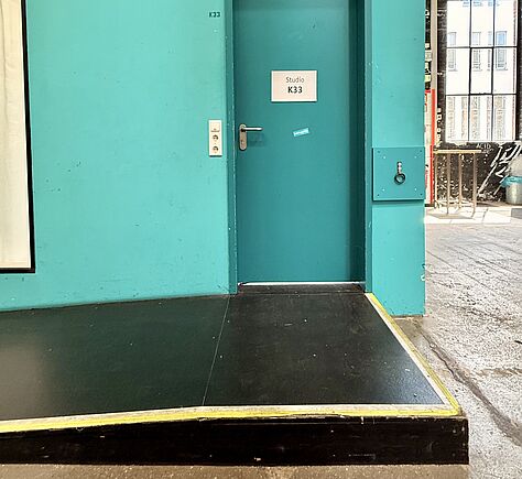 Auf einer türkisfarbenen Tür ist ein weißes Schild mit der Aufschrift 'Studio K33’. Die Tür befindet sich in einer türkis gestrichenen Wand. Vor der Tür führt von links eine schwarze Rampe mit gelbem Randstreifen nach oben. Die Rampe ist etwa eine Stufe hoch. Links neben der Tür ist ein Lichtschalter und darunter zwei ungeschützte Steckdosen. Rechts ist eine türkisfarbene Platte mit einem kleinen Metallring. Der Boden besteht aus grauem Beton, im Hintergrund ist ein großes Fenster.